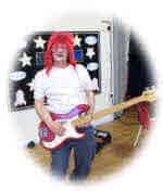 Adrian with a red wig plays a bass guitar.